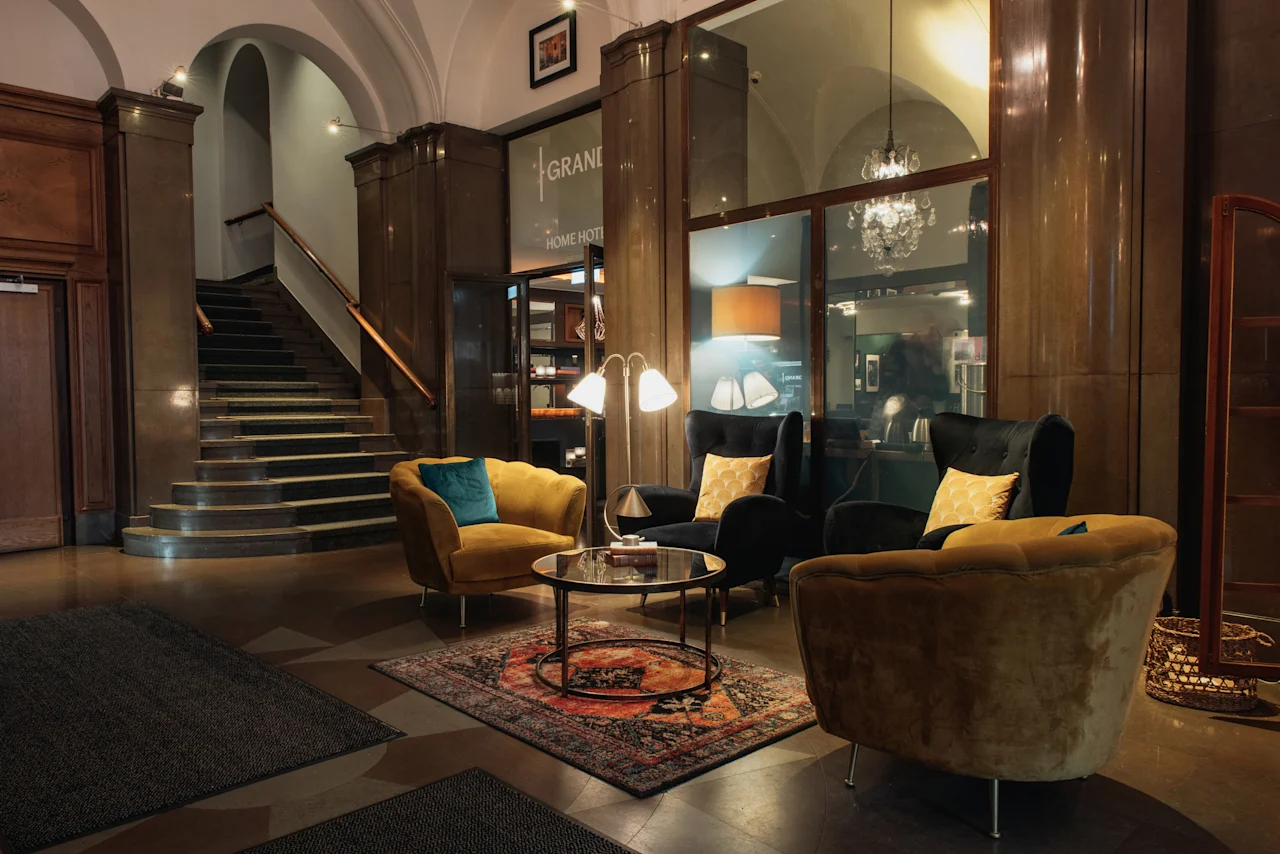 En välkomnande hotellobby på Home Hotel Grand, Helsingborg, med en storslagen trappa, plyschmöbler och mjuk belysning. Text: GRAND HOME HOTEL.