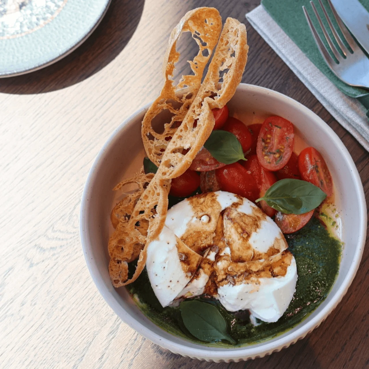 A bowl of fresh burrata cheese drizzled with balsamic glaze sits with cherry tomatoes, basil, and crispy bread on a wooden table. Nearby, a green napkin holds cutlery with a logo.