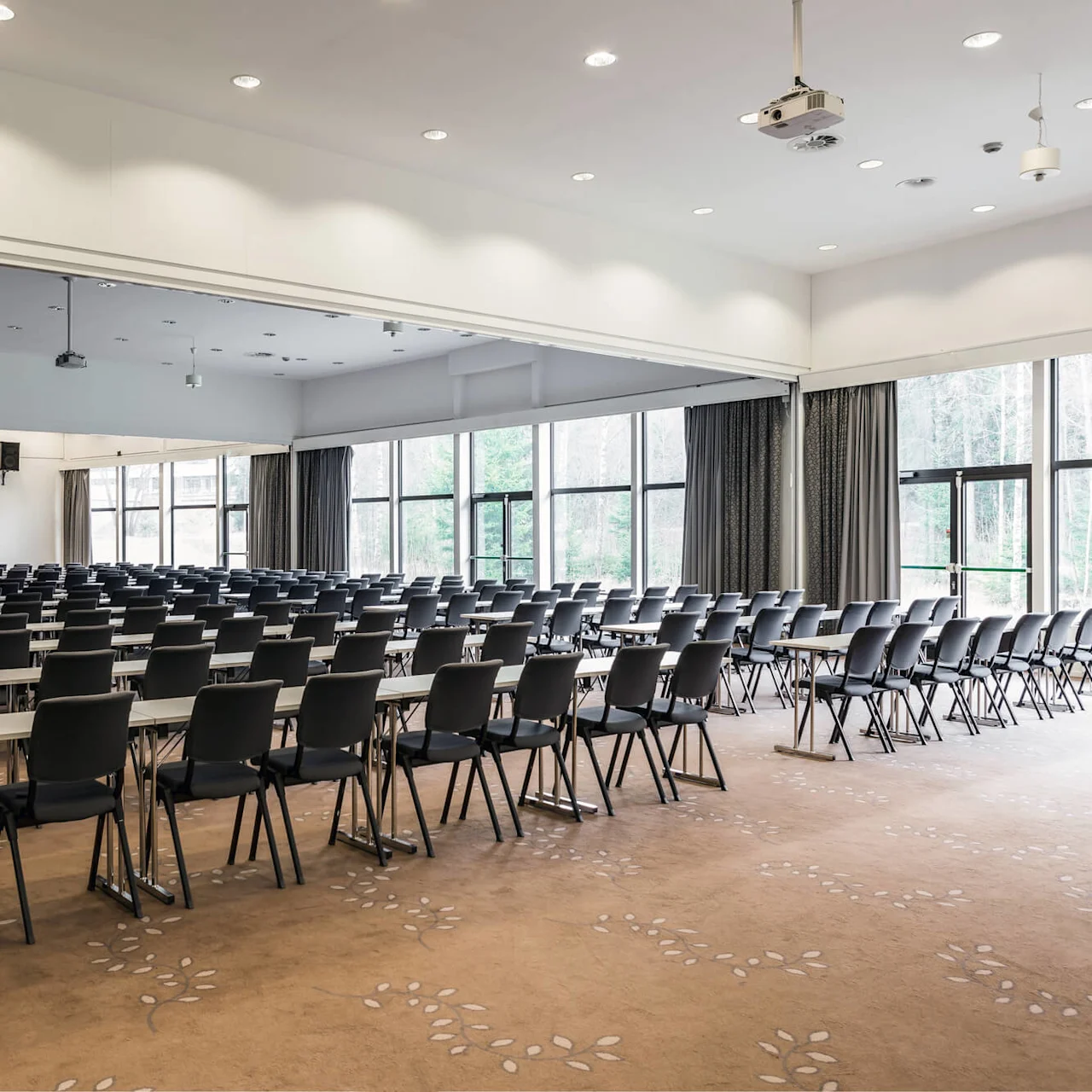Rows of black chairs and tables face a large white projector screen in a spacious conference room with beige carpeting and large windows.