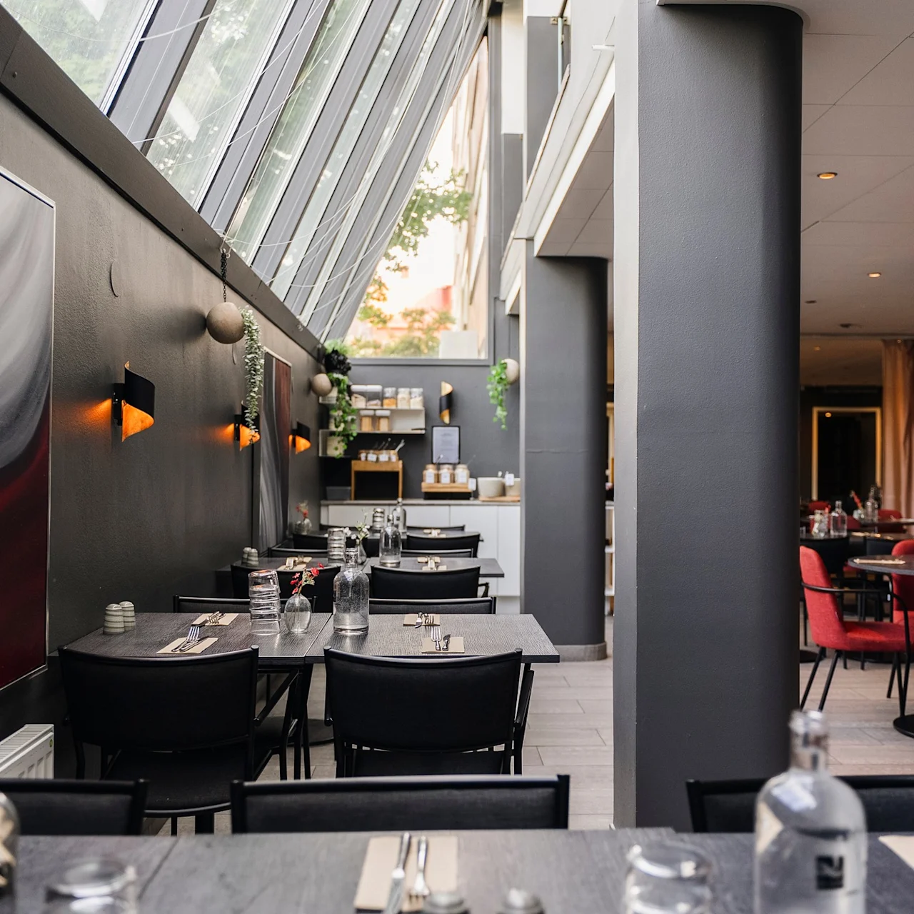 Samtida restauranginteriör med prydligt dukade bord med svarta stolar, grå väggar prydda med abstrakt konst och ett stort snedställt fönster som släpper in naturligt ljus. Röda stolar fyller den rymliga matsalen.