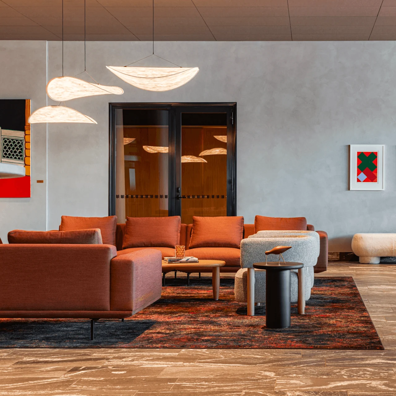 A seating area features orange and grey sofas with cushions and coffee tables on a patterned rug. Overhead, modern pendant lamps illuminate, with abstract artwork on the wall.