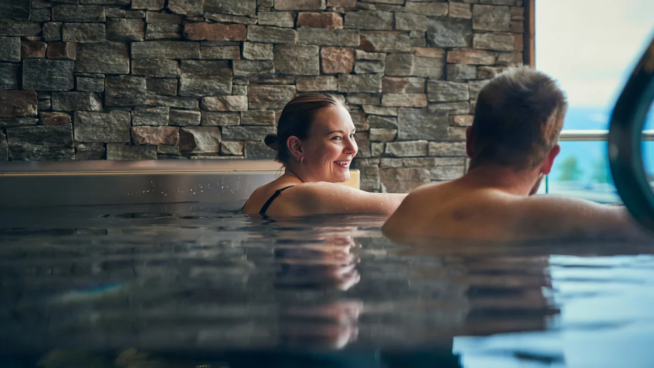 A woman and a man relax in a spa pool, smiling and engaging in conversation. They are surrounded by stone walls, creating a warm, serene atmosphere. 