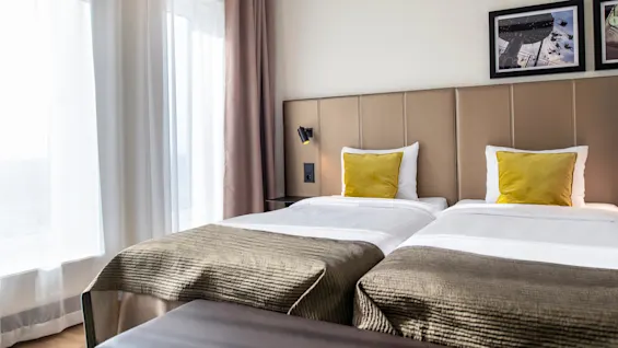 Two neatly made twin beds with yellow cushions, under framed artwork, situated in a warmly lit room with beige curtains covering large windows.