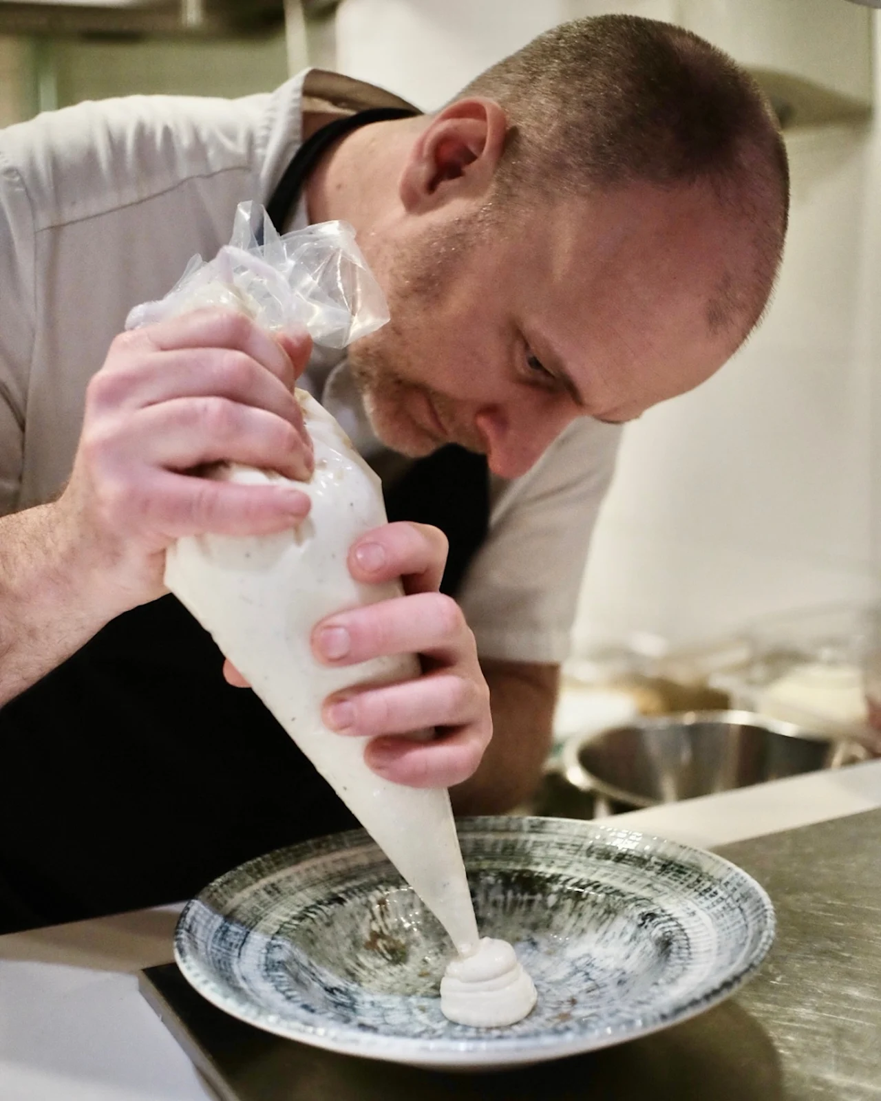 A chef concentrates while using a piping bag to shape cream onto a decorative plate in a professional kitchen.