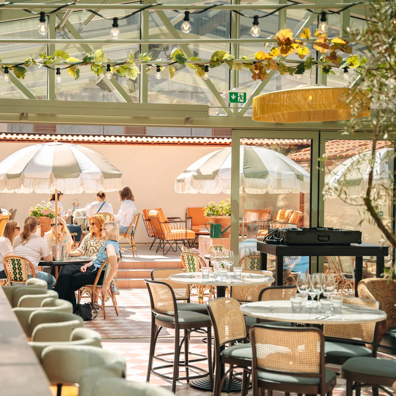 People sit at tables under large umbrellas on a sunny patio, surrounded by potted plants and soft lighting. A bar counter is visible on the left.