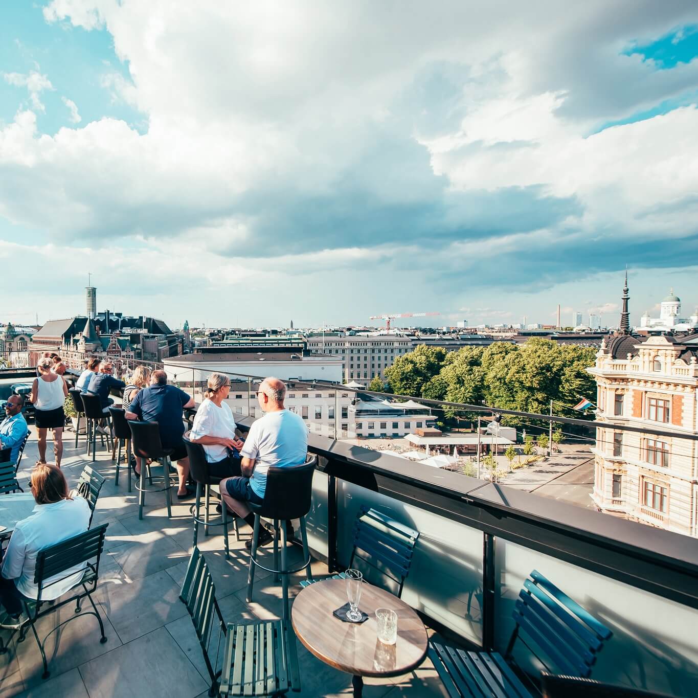 Ihmisiä seurustelee kattoterassibaarissa kaupunkimaiseman äärellä kirkkaan taivaan alla.