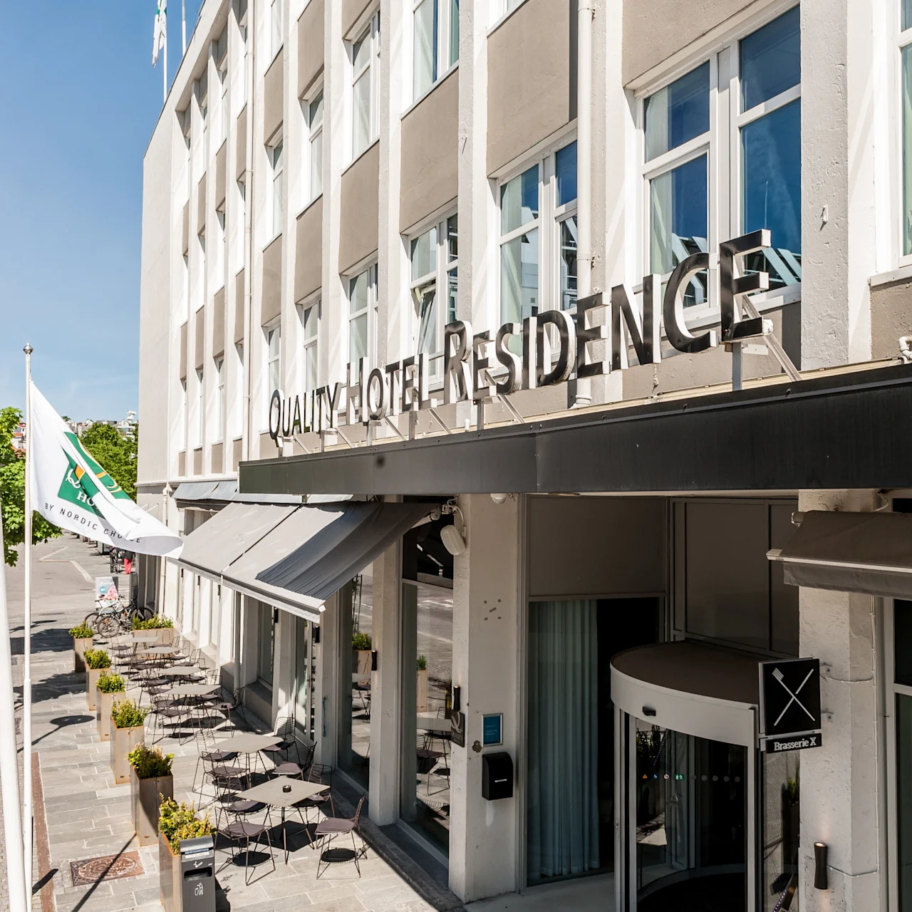 Hotellin sisäänkäynti, jonka yläpuolella on kyltti "Quality Hotel Residence", ulkona istumapaikkoja ja istutuksia. Lippuja ja pyöröovi. Kirkas, aurinkoinen päivä.