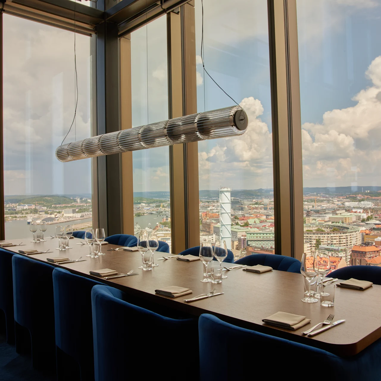 Ett långt träbord med blå stolar är dukat med glas och bestick, placerat i ett höghusrum med stora fönster som vetter mot en stadsbild under en molnig himmel.