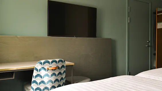 Et vægmonteret TV hænger over et skrivebord og en mønstret stol i et ryddeligt, moderne rum med en seng og en lukket dør.