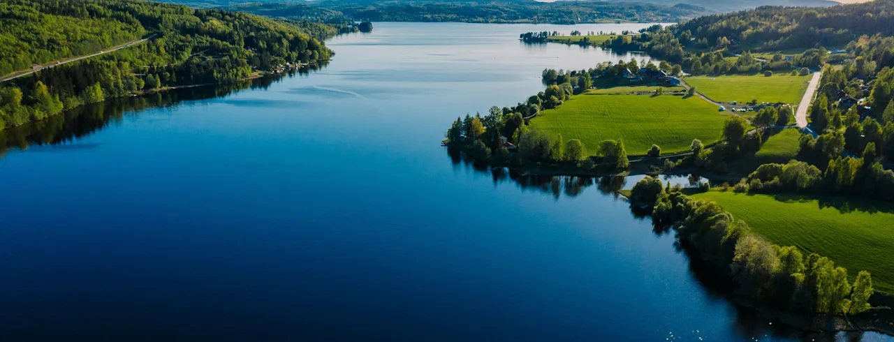 Flygfoto över en lugn sjö omgiven av frodiga gröna skogar och fält under en klarblå himmel.