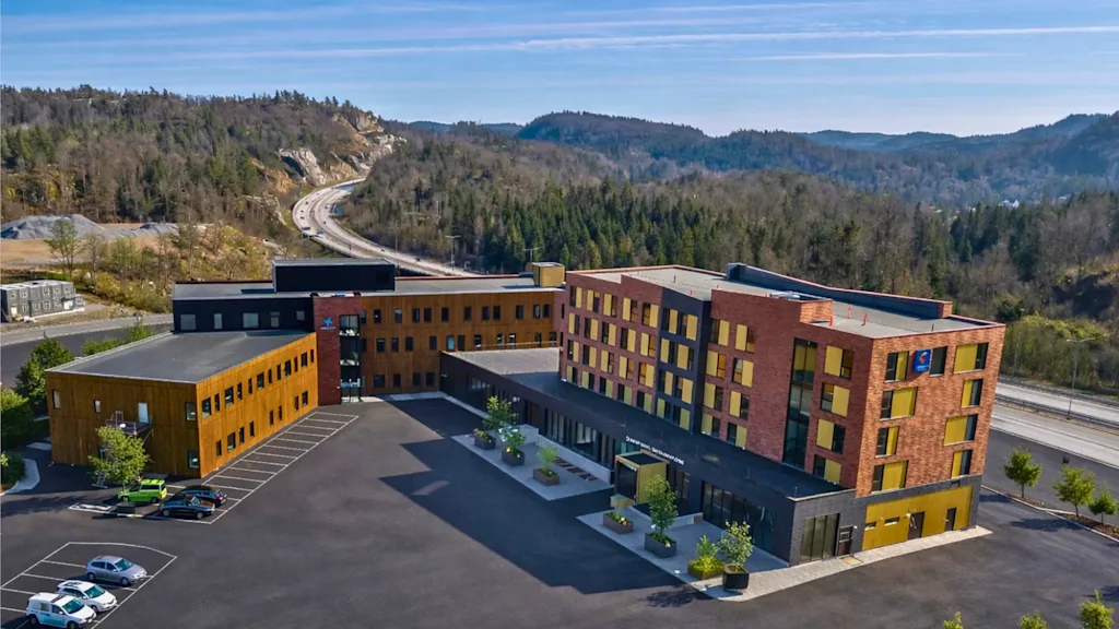 En moderne hotellbygning med en stor parkeringsplass, mot et bakteppe av skogkledde åser og en svingete vei.