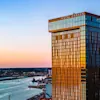 A modern hotel building with "HOTEL DRAKEN" sign, reflecting sunset hues, overlooking a city and a body of water.