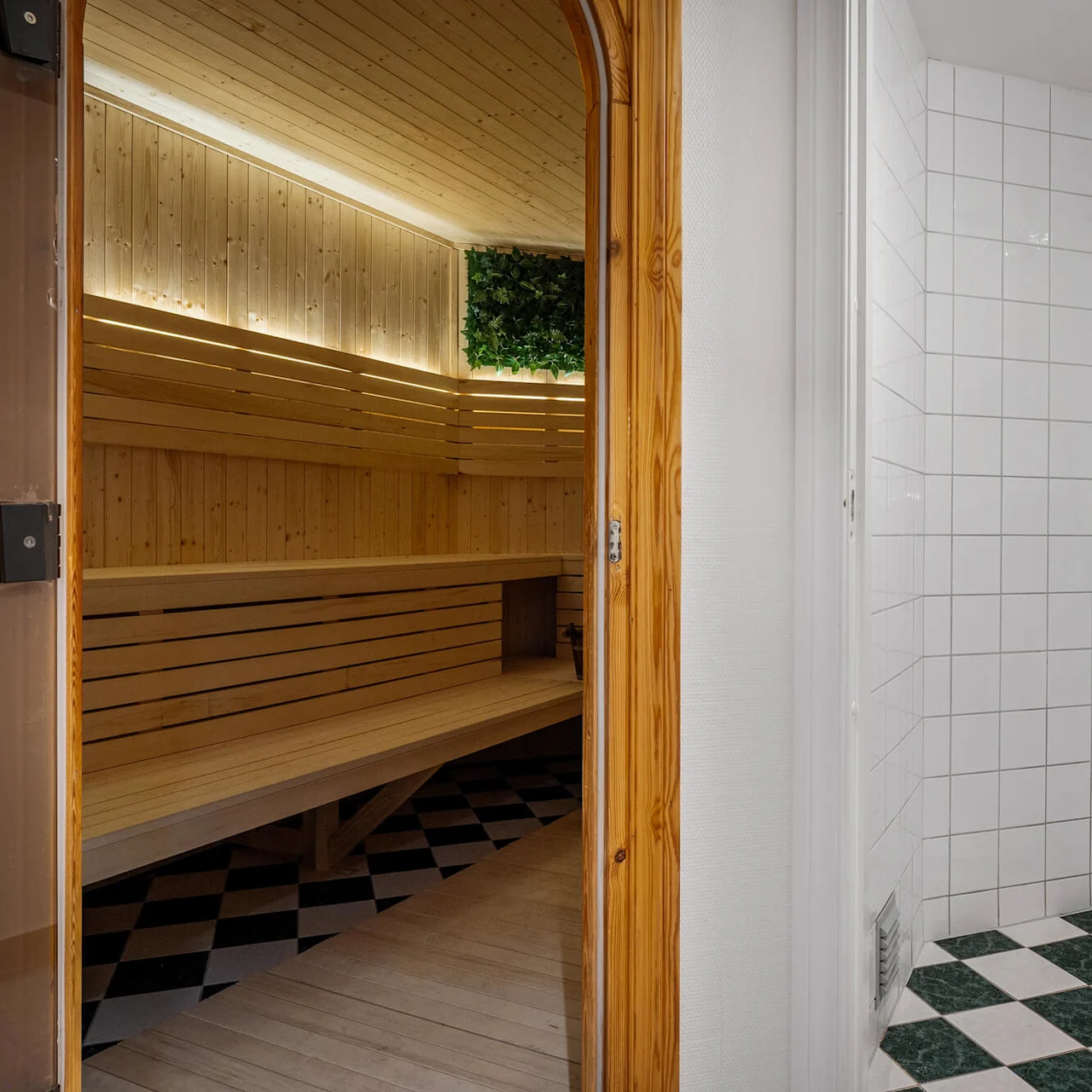 Sauna med træbænke er synlig gennem en åben trødør, ved siden af en hvidflisebelagt bruser med et sølvfarvet brusehoved.