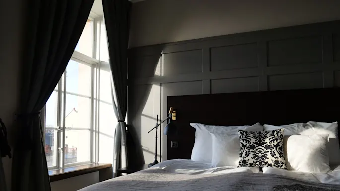 Seng med hvite og mønstrede puter i mykt lys. Sollys strømmer gjennom store vinduer med mørke gardiner, som lyser opp det koselige soverommet.