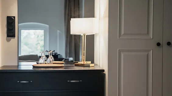 Et koselig hotellrominteriør med en kommode, en lampe og vinglass, som reflekterer et vindu. Home Hotel Norre Park i Halmstad.