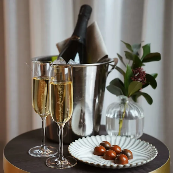 Två champagneglas med bubblor bredvid en ishink med en flaska, på ett bord med choklad, intill gardiner.