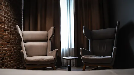 Two armchairs sit by a small round table in front of a window with dark curtains, set against a brick wall backdrop.