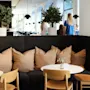 Comfortable seating area with beige cushions and a small round table, surrounded by wooden chairs. Two potted plants are placed on the partition. People stand near large windows in the background.