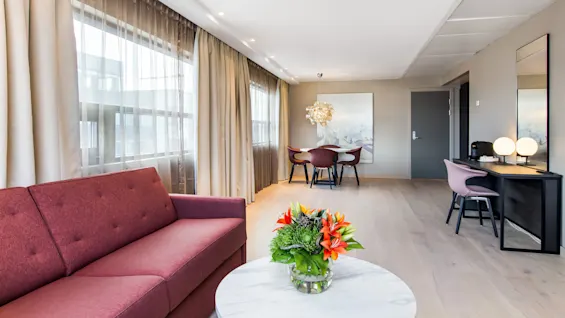 A maroon sofa sits beside a round table with vibrant flowers in a bright room. Large windows are covered with sheer curtains. The background reveals a dining area.