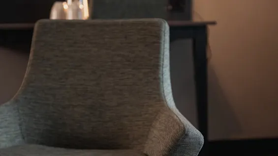 Viihtyisä hotellihuoneen nurkkaus, jossa on mukava harmaa tuoli, tumma pöytä ja pehmeä valaistus seinävalaisimesta.
