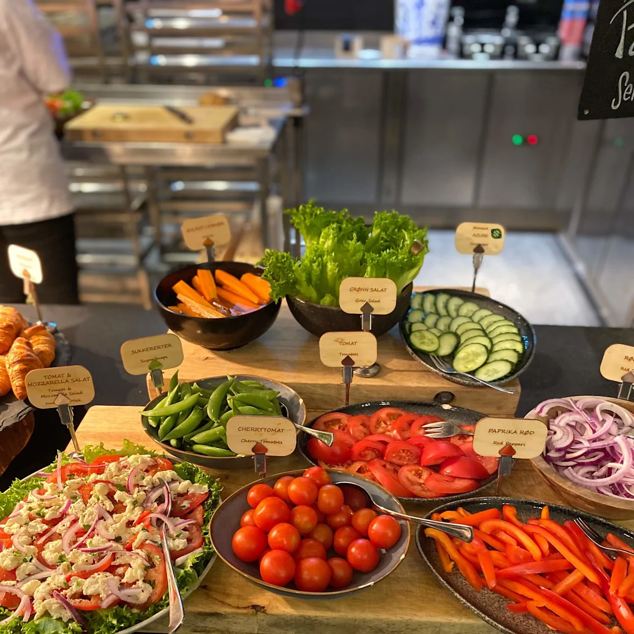 A buffet with assorted fresh vegetables is arranged on platters with labels including "Tomat & Mozzarella Salat," "Sukkererter," "Gulerødder," "Grønn Salat," "Cherrytomat," "Paprika Rød." Behind is a kitchen setting.