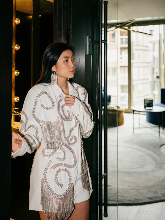 A person in a dazzling white jacket with sparkling fringe stands in a doorway, looking out into a bright room with a city view.