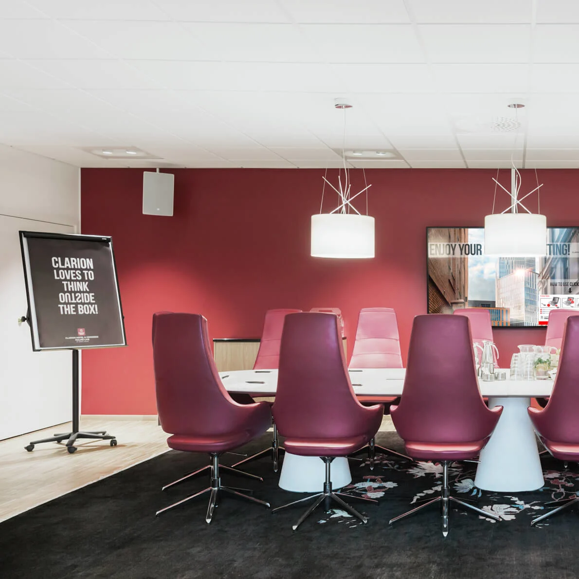 Et møterom har et langt bord omgitt av røde stoler. Et skilt lyder "CLARION LOVES TO THINK OUTSIDE THE BOX," mot en vinrød vegg med moderne belysning.