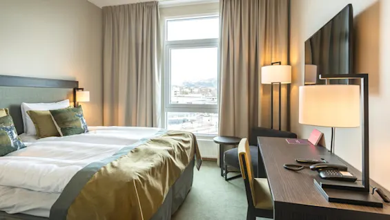 Siististi pedattu sänky valoisassa hotellihuoneessa, moderneja huonekaluja, isot ikkunat ja näkymä kaupunkiin.