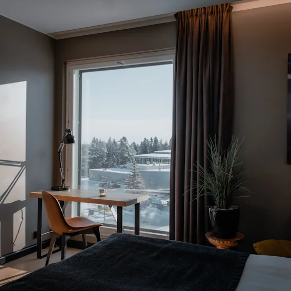 Hotellihuone, jossa työpöytä suuren ikkunan ääressä, näkymä lumiseen maisemaan ja puihin. Viihtyisä ja valoisa tila.