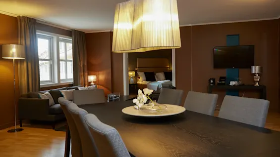 A dining table adorned with a floral centerpiece sits under a modern pendant light. Nearby are a cozy seating area, a bed visible through a doorway, and a wall-mounted TV.