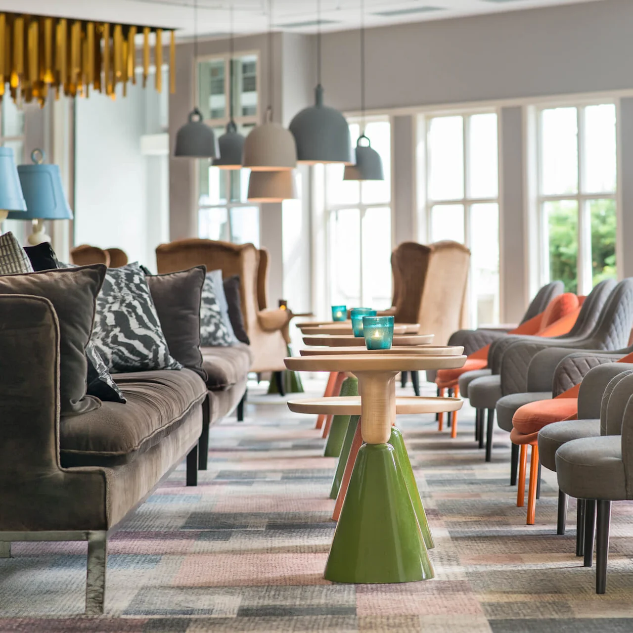 Modern lounge chairs and sofas are arranged symmetrically around wooden tables with blue candles, under hanging lamps, in a bright room with large windows overlooking greenery.