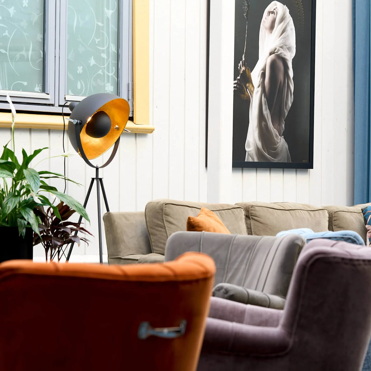 En hyggelig stue med forskellige sofaer, en gulvlampe, planter og et stort indrammet kunstværk på væggen.