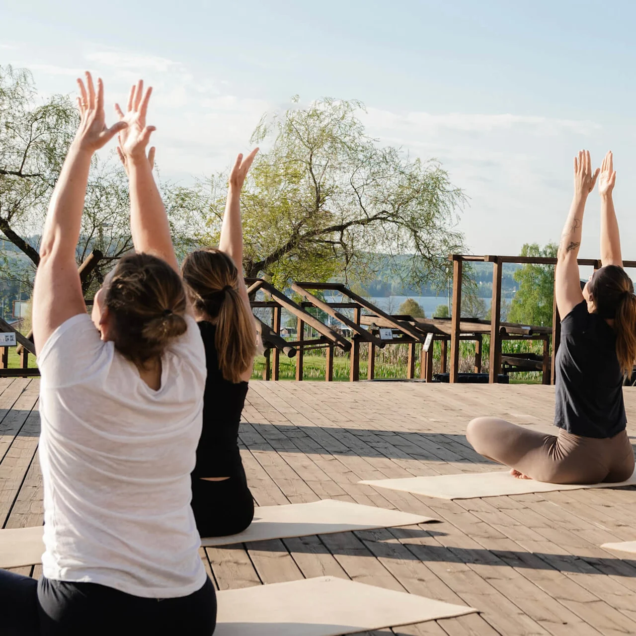 Personer sitter på yogamatter, med armene hevet, og praktiserer yoga utendørs på en tredekke. Trær og en innsjø er synlige i bakgrunnen.