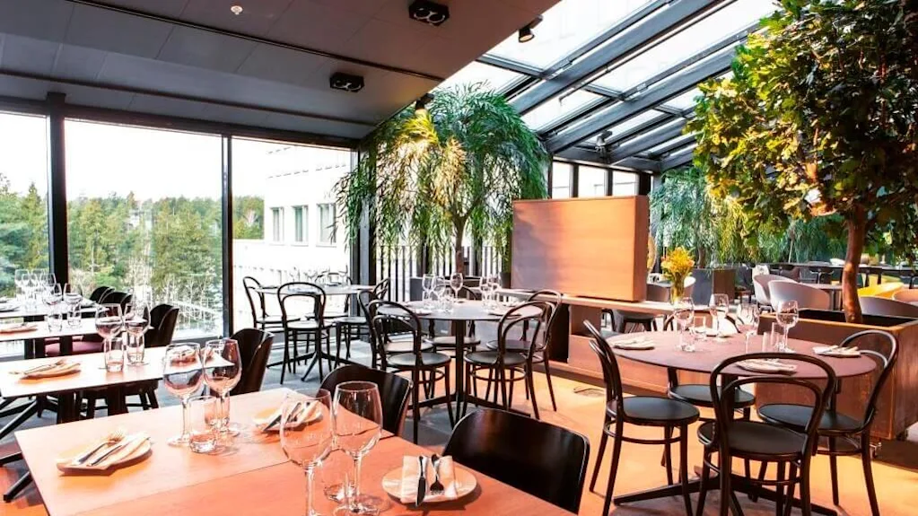 Restaurangbord är dukade med glas och bestick under ett glastak. Gröna växter omger sittgruppen, och stora fönster erbjuder en utsikt över ett skogsområde.