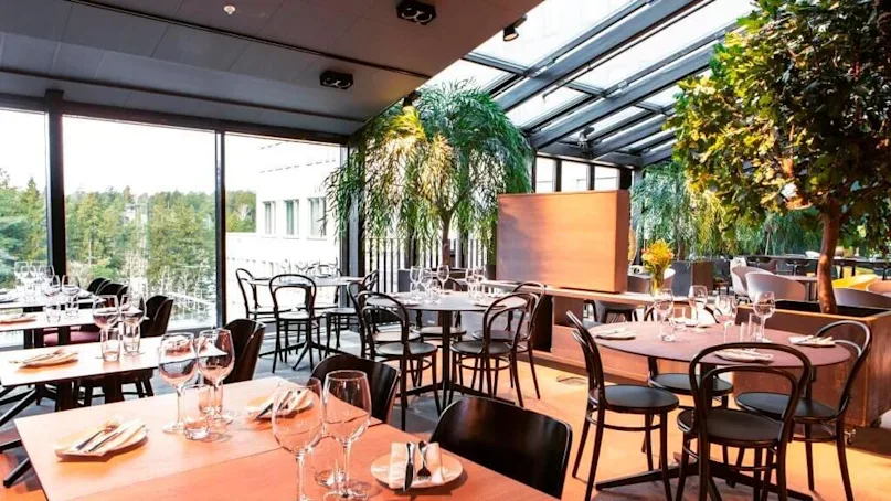Restaurant tables are set with glasses and utensils under a glass ceiling. Green plants surround the seating area, and large windows offer a view of a forested landscape.