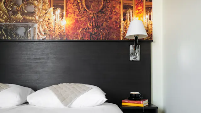 Bed with white pillows sits under dark wood headboard; a wall-mounted light fixture is above a bedside table holding books and a black coffee cup. Ornate art decorates the wall.