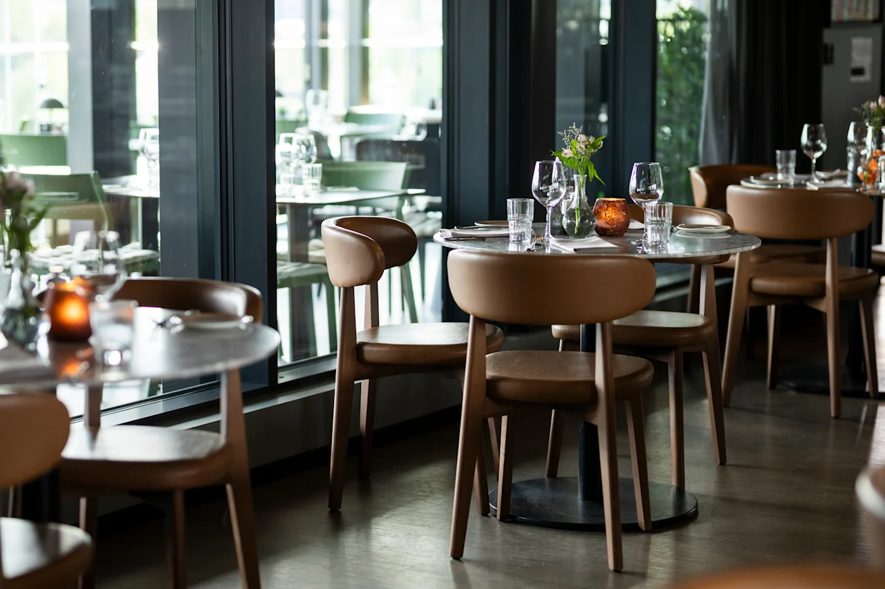 Et koselig restaurantinteriør med moderne brune stoler og runde bord dekket med glass, tallerkener, blomster og tente lys, ved store vinduer.