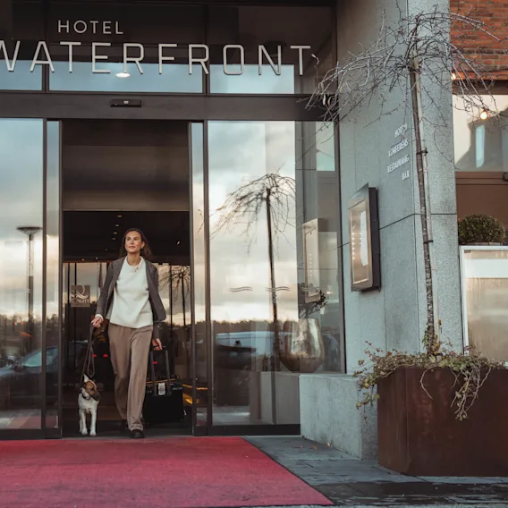 En kvinna går ut från "Hotel Waterfront" med en resväska och en hund i koppel. Glasentrén speglar utsidan, kantad med växter och en röd matta.
