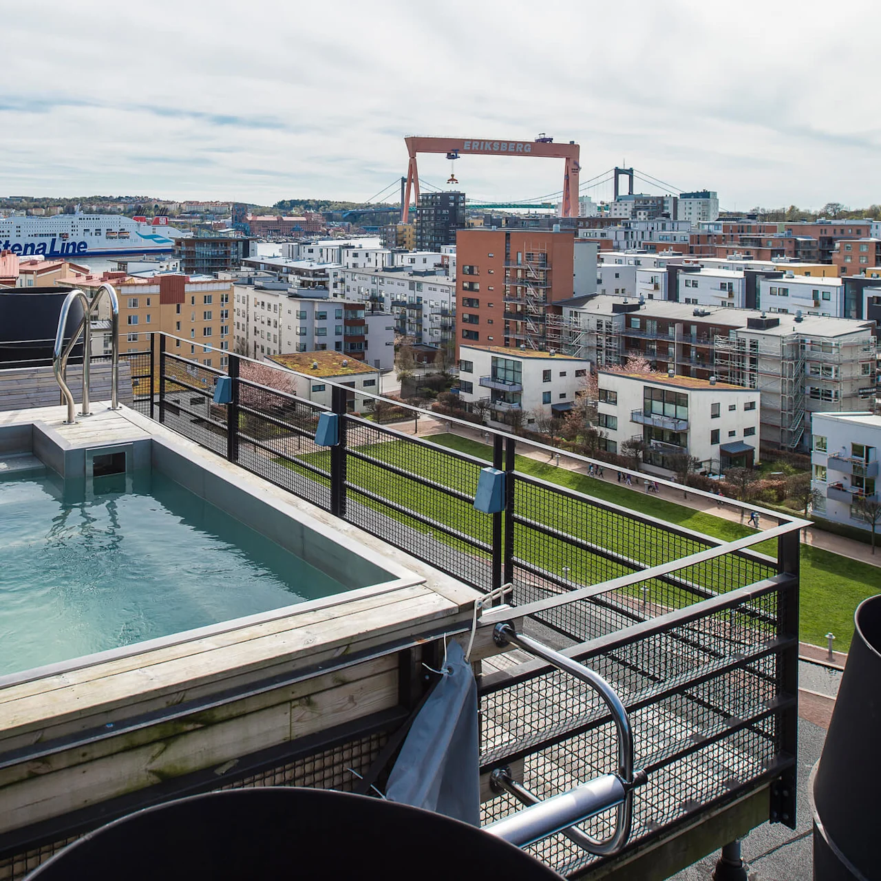 A rooftop pool with a metal ladder overlooks a cityscape, including residential buildings, a green park, and a large crane labeled "ERIKSBERG." A ferry with "Stena Line" visible in the background.