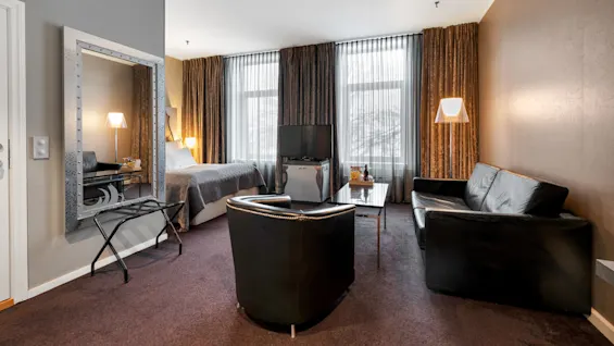 Et hotelværelse med dobbeltseng, lædersofa og fjernsyn, med gulv-til-loft-vinduer og brune gardiner, skaber en hyggelig og moderne atmosfære.
