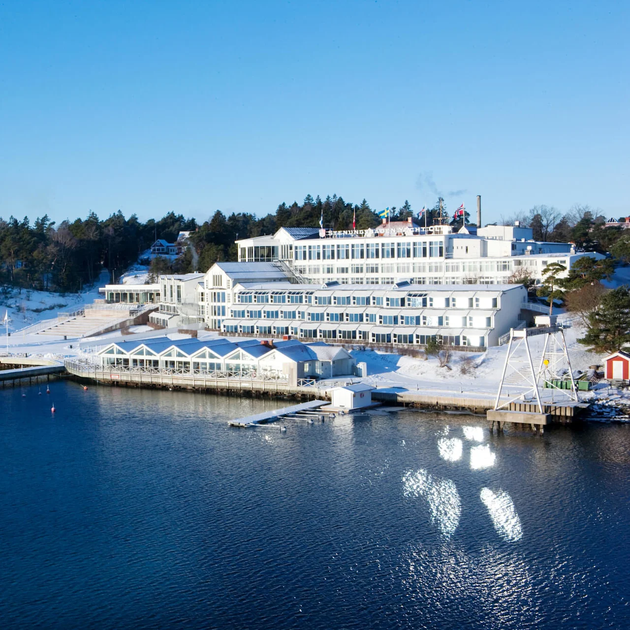 A large white building with the text "Stenungsbaden Yacht Club" stands near a snow-covered shoreline by the water, surrounded by trees under a clear blue sky.