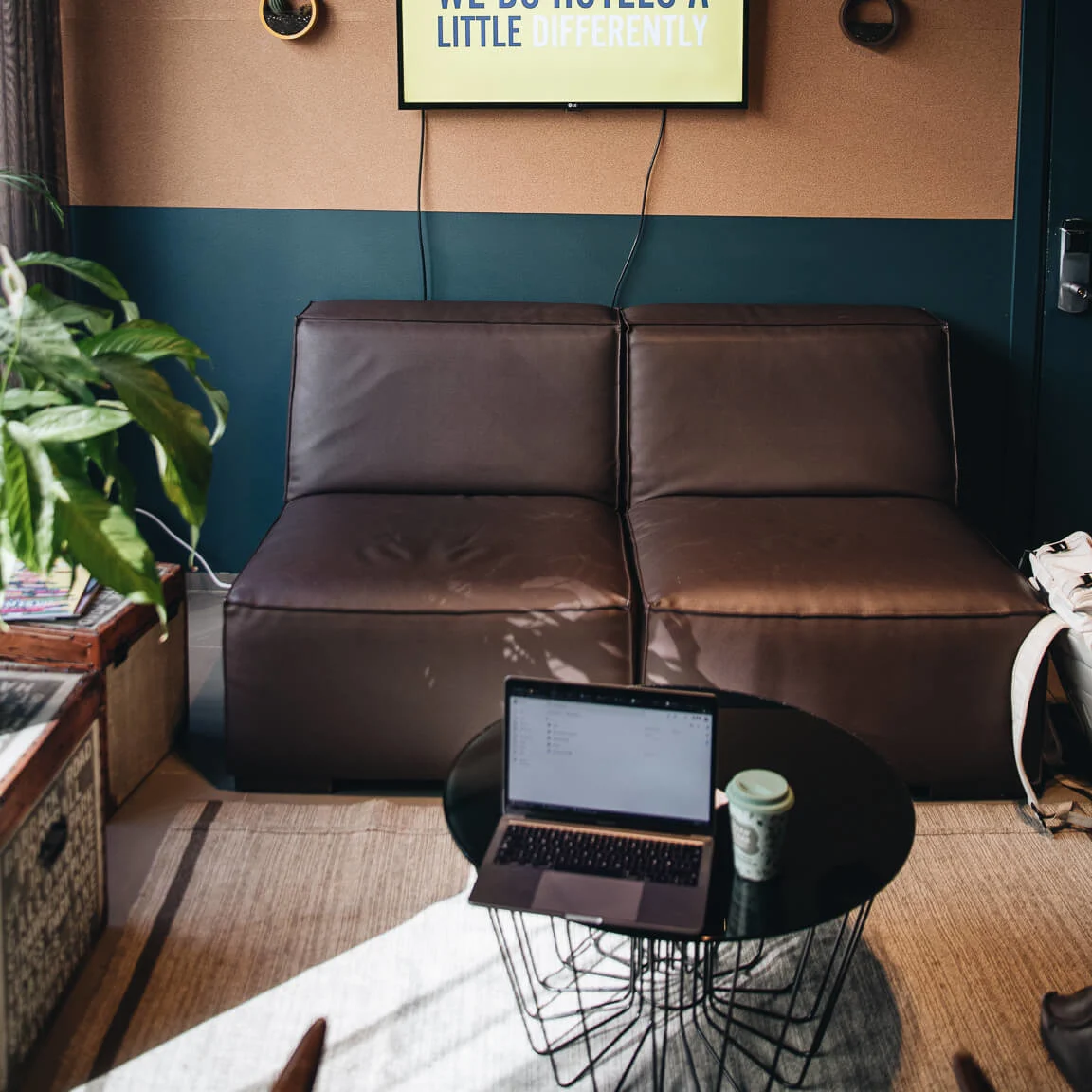 Television displaying "WE DO HOTELS A LITTLE DIFFERENTLY" hangs above a brown couch. A coffee table with a laptop and cup in a modern, cozy room with plants.
