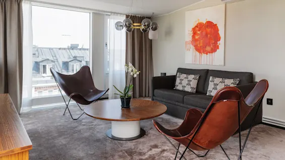 Ett modernt vardagsrum med stilfulla möbler och ett stort konstverk, med utsikt över stadstak genom ett fönster.