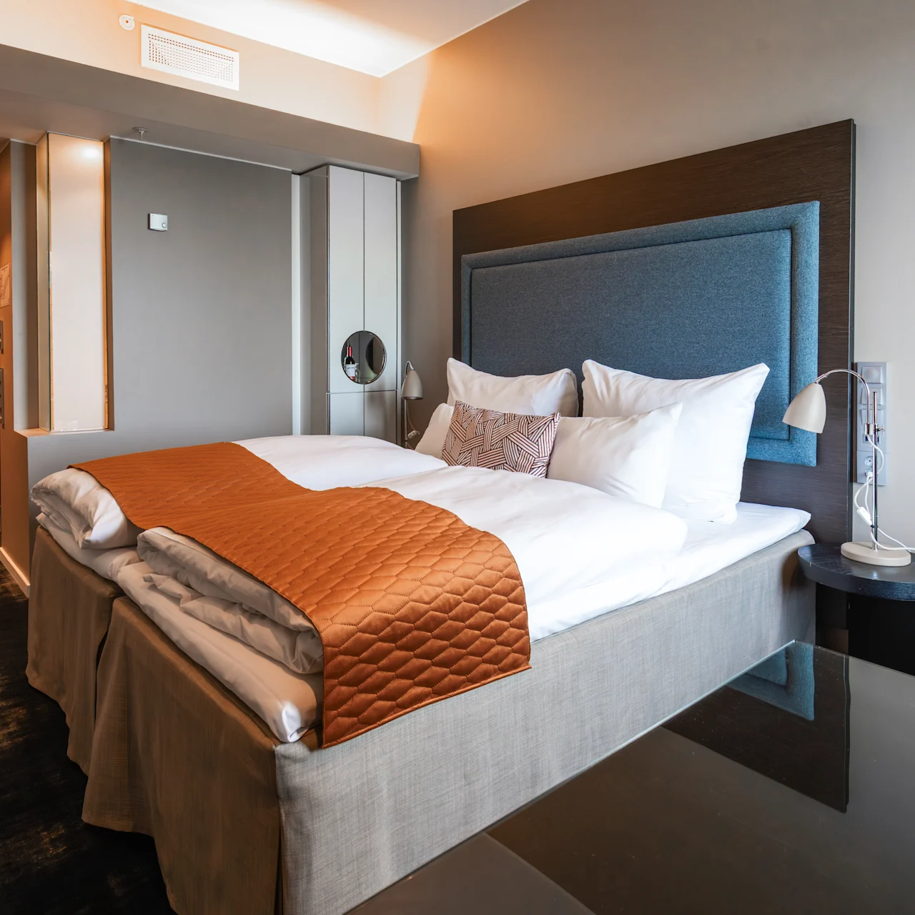 A neatly arranged bed with white linens and an orange quilt lies in a modern hotel room. Nearby, a turquoise chair and dark wooden door complement the neutral-toned decor.