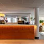 Reception med datorer och en person bakom. Neonljusskylt lyder "BOKA RUM." Modern lounge med soffor och växter. Stora fönster släpper in naturligt ljus.