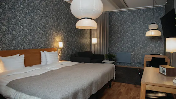A hotel room with two beds, covered with a beige blanket, stands in a modern, wallpapered room. A desk, lamps, and a small couch complete the cozy setting.