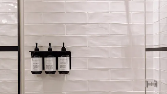Three bottles with pumps are mounted on a white tiled shower wall. They are labelled "SPREKENHUS" with products: "BODY WASH," "HYDRATING SHAMPOO," and "HYDRATING CONDITIONER." 