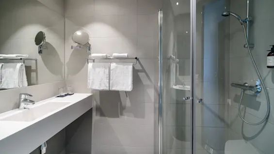 Sleek bathroom; white sink beside a glass shower enclosure beneath bright lighting; modern fixtures and neat towels create a clean, minimalist ambiance.