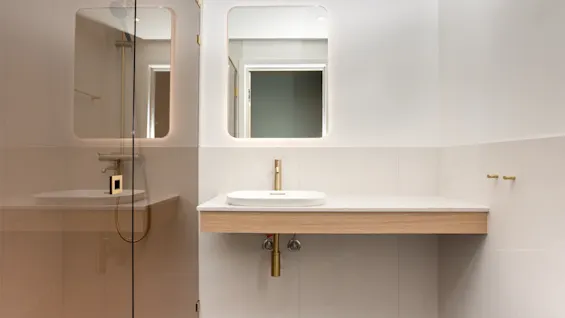 Et moderne badeværelse med et stilrent vaskemøbel, oplyst spejl og en bruser med glasvæg, med guldfarvede armaturer.