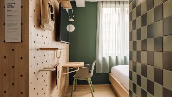 A modern hotel room with a wooden pegboard wall, desk, chair, and bed. 